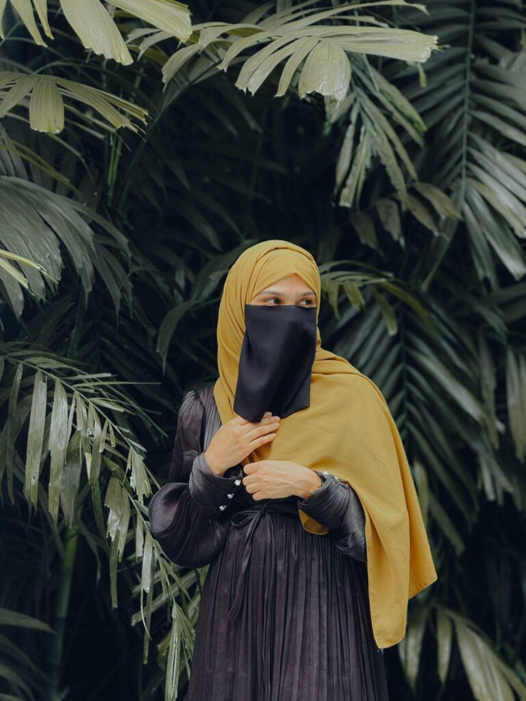 A freelance digital marketing consultant in malappuram woman ,wearing a yellow hijab and black niqab, dressed in a dark pleated outfit, standing in front of lush green tropical leaves.
